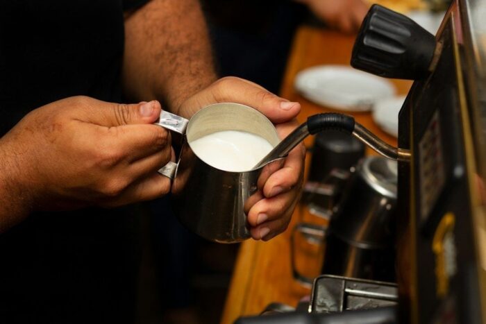 Hände halten ein Milchkännchen unter die Dampfdüse einer Espressomaschine